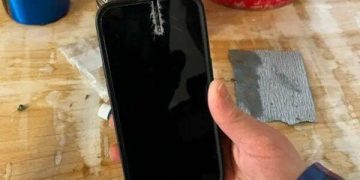 Hand holding damaged iPhone with saw cut in a workshop setting.