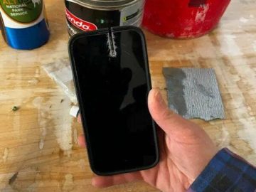 Hand holding damaged iPhone with saw cut in a workshop setting.