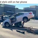 Car stuck on pole near shopping center, driver still on phone.