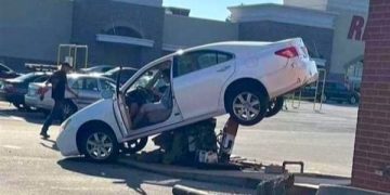Car stuck on pole near shopping center, driver still on phone.