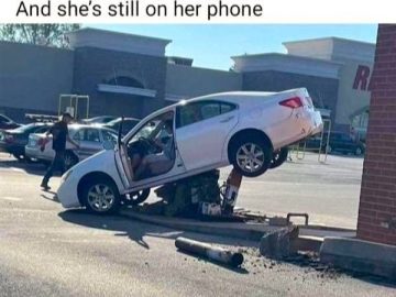 Car stuck on pole near shopping center, driver still on phone.