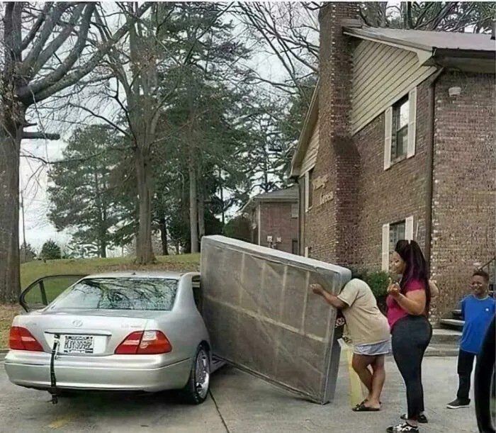 Three people struggling to fit a mattress into a car.