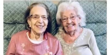Two elderly women smiling, holding hands in a retirement home.