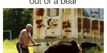Man confidently chases a bear with a stick in front of a trailer.