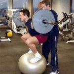 Man squatting on a gym ball with weights, about to fall