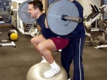 Man squatting on a gym ball with weights, about to fall