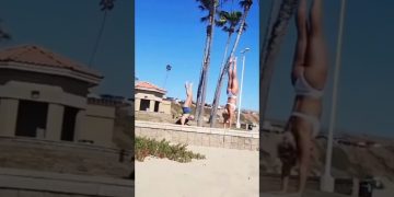 A woman attempting a handstand on a beach wall loses balance and falls into the sand.