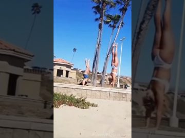 A woman attempting a handstand on a beach wall loses balance and falls into the sand.