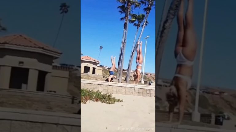 A woman attempting a handstand on a beach wall loses balance and falls into the sand.