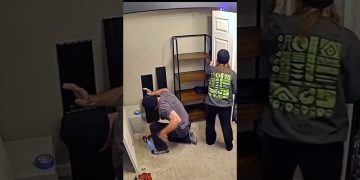 A woman carrying a long white board accidentally bumps into a kneeling man during a DIY shelf project.